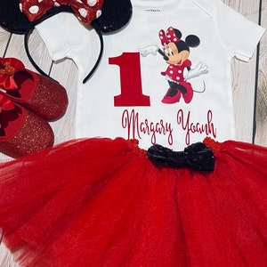 May include: Red and black Minnie Mouse themed first birthday outfit. The outfit includes a white bodysuit with a red number one, a red tulle tutu, a black bow, and red glitter shoes with a Minnie Mouse bow.