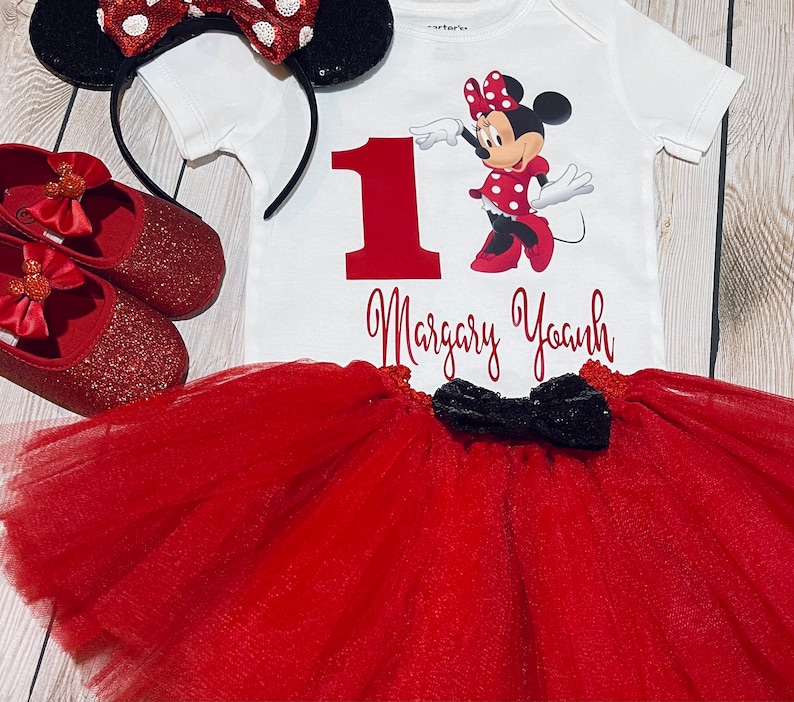 May include: Red and black Minnie Mouse themed first birthday outfit. The outfit includes a white bodysuit with a red number one, a red tulle tutu, a black bow, and red glitter shoes with a Minnie Mouse bow.