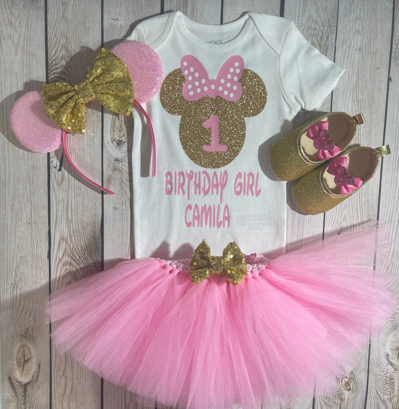 May include: A pink and gold Minnie Mouse themed first birthday outfit for a baby girl. The outfit includes a white bodysuit with a gold glitter Minnie Mouse head and the text "1 Birthday Girl Camila", a pink tulle tutu with a gold bow, a pink headband with gold glitter ears, and a pair of gold glitter shoes with pink bows.