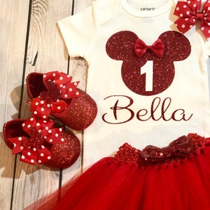 Puede incluir: Conjunto de primer cumplea&ntilde;os de Minnie Mouse rojo y blanco para una ni&ntilde;a. El conjunto incluye un body blanco con una cabeza de Minnie Mouse roja de purpurina, el n&uacute;mero uno y el nombre Bella. El conjunto tambi&eacute;n incluye una falda de tul roja con un lazo rojo de purpurina y zapatos rojos de purpurina con lazos de lunares rojos y blancos.