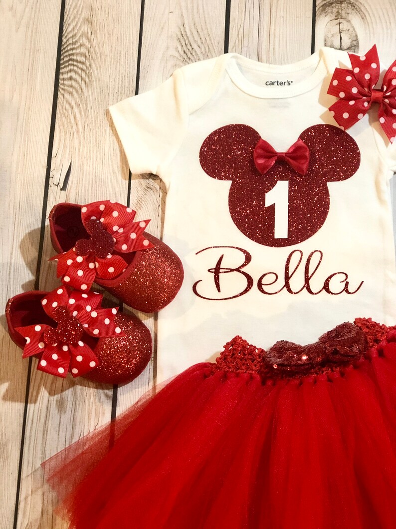 Puede incluir: Conjunto de primer cumplea&ntilde;os de Minnie Mouse rojo y blanco para una ni&ntilde;a. El conjunto incluye un body blanco con una cabeza de Minnie Mouse roja de purpurina, el n&uacute;mero uno y el nombre Bella. El conjunto tambi&eacute;n incluye una falda de tul roja con un lazo rojo de purpurina y zapatos rojos de purpurina con lazos de lunares rojos y blancos.