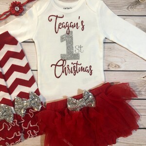 May include: A white long-sleeved onesie with the text "Teagan's 1st Christmas" in red glitter. The outfit includes red and white chevron leggings, a red tutu skirt, and a red headband with a flower. Silver sequin bows adorn the skirt and leggings.
