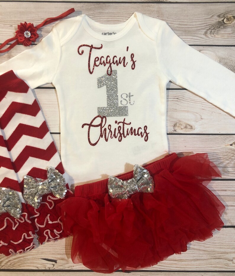 May include: A white long-sleeved onesie with the text "Teagan's 1st Christmas" in red glitter. The outfit includes red and white chevron leggings, a red tutu skirt, and a red headband with a flower. Silver sequin bows adorn the skirt and leggings.