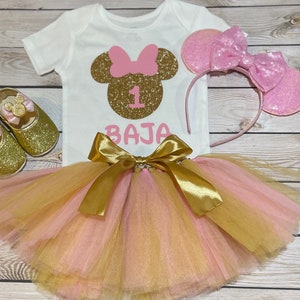 May include: A pink and gold Minnie Mouse themed first birthday outfit for a baby girl. The outfit includes a white bodysuit with a gold glitter Minnie Mouse head, a pink bow, and the number 1. The outfit also includes a pink and gold tulle tutu with a gold satin bow and a pink headband with glitter Minnie Mouse ears.