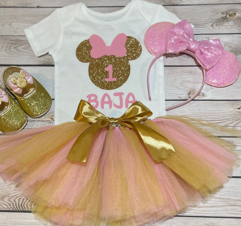 May include: A pink and gold Minnie Mouse themed first birthday outfit for a baby girl. The outfit includes a white bodysuit with a gold glitter Minnie Mouse head, a pink bow, and the number 1. The outfit also includes a pink and gold tulle tutu with a gold satin bow and a pink headband with glitter Minnie Mouse ears.