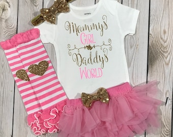 May include: A white onesie with the text "Mommy's Girl Daddy's World" in pink and gold glitter. The onesie is paired with a pink tulle skirt with a gold glitter bow, pink and white striped leg warmers with gold glitter hearts, and a gold glitter headband with a bow.
