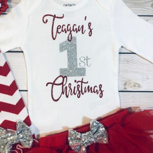 May include: A white baby bodysuit with the text "Teagan's 1st Christmas" in red glitter and silver glitter. The outfit includes a red tutu skirt with silver sequin bows, red and white striped leggings, and a red headband.