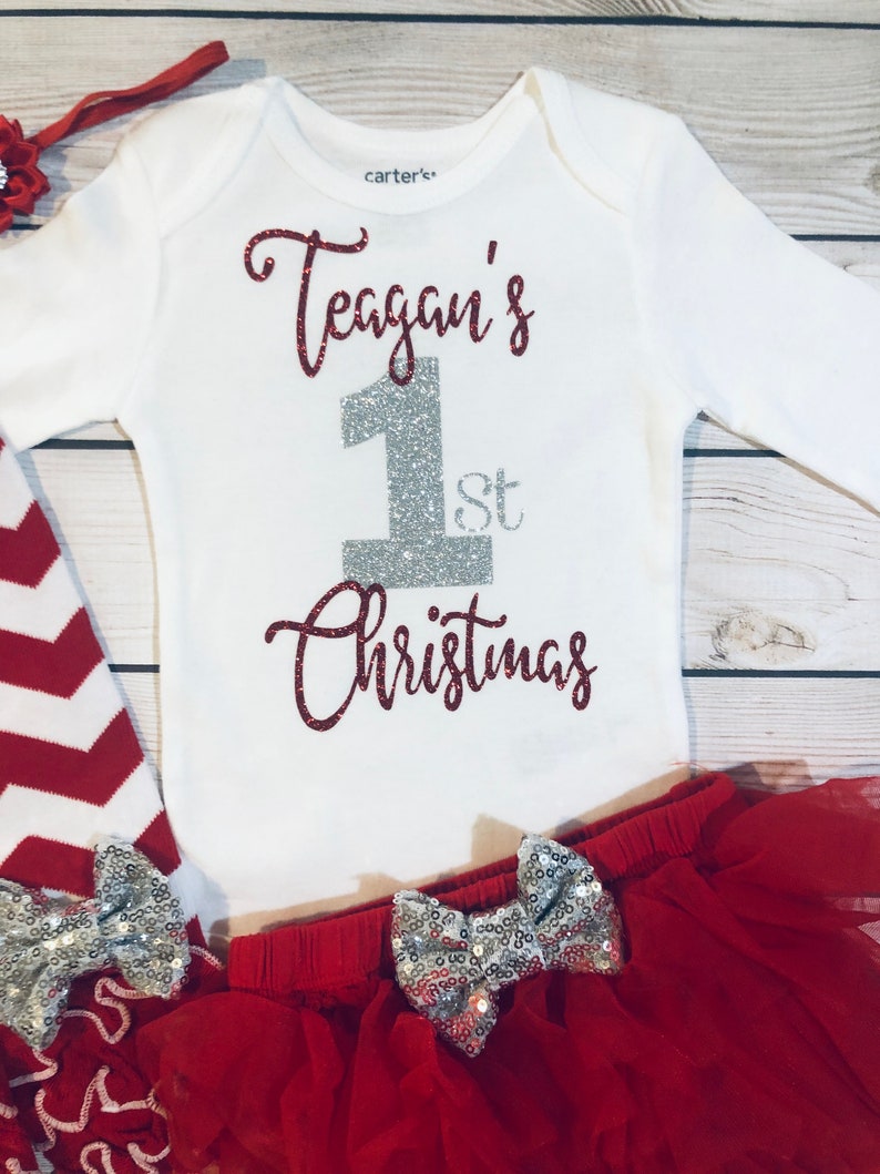 May include: A white baby bodysuit with the text "Teagan's 1st Christmas" in red glitter and silver glitter. The outfit includes a red tutu skirt with silver sequin bows, red and white striped leggings, and a red headband.