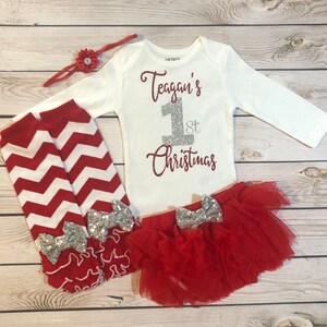 May include: A white baby bodysuit with the text "Teagan's 1st Christmas" in red and silver glitter. The set includes a red tutu skirt, leg warmers with silver bows, and a red headband with a flower.