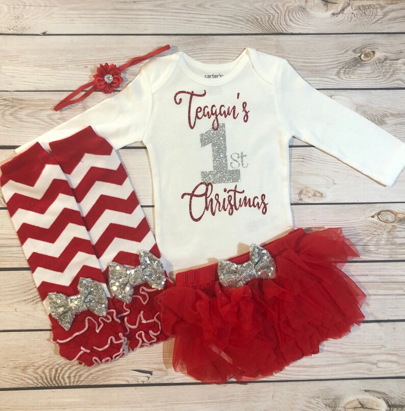 May include: A white baby bodysuit with the text "Teagan's 1st Christmas" in red and silver glitter. The set includes a red tutu skirt, leg warmers with silver bows, and a red headband with a flower.