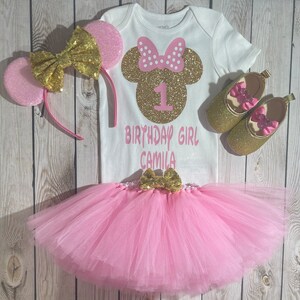 May include: A pink and gold first birthday outfit for a girl. The outfit includes a white onesie with a gold glitter Minnie Mouse head, the number 1, and the text "Birthday Girl Camila". The outfit also includes a pink tulle tutu, a pink and gold headband with Minnie Mouse ears, and a pair of gold glitter shoes with pink bows.