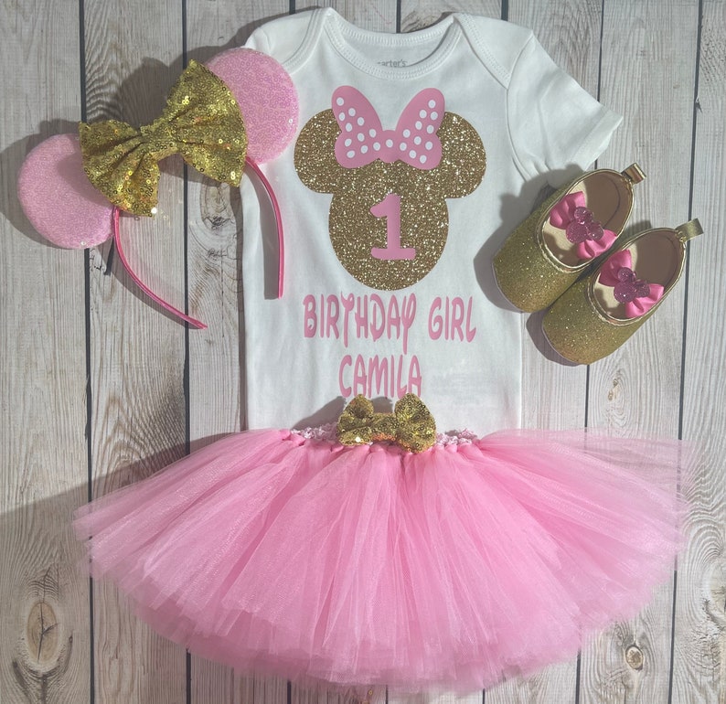 May include: A pink and gold first birthday outfit for a girl. The outfit includes a white onesie with a gold glitter Minnie Mouse head, the number 1, and the text "Birthday Girl Camila". The outfit also includes a pink tulle tutu, a pink and gold headband with Minnie Mouse ears, and a pair of gold glitter shoes with pink bows.