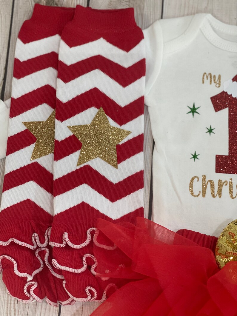 Pu&ograve; includere: Un completo festivo per neonati con scaldamuscoli a zig-zag rossi e bianchi con stelle glitterate dorate, un body bianco con "My 1st Christmas" in glitter dorati e un tut&ugrave; rosso. Il completo &egrave; pensato per il primo Natale di un bambino.