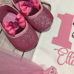 May include: Pink glitter baby shoes with a pink bow and a Minnie Mouse head design. The shoes are on a wooden surface.