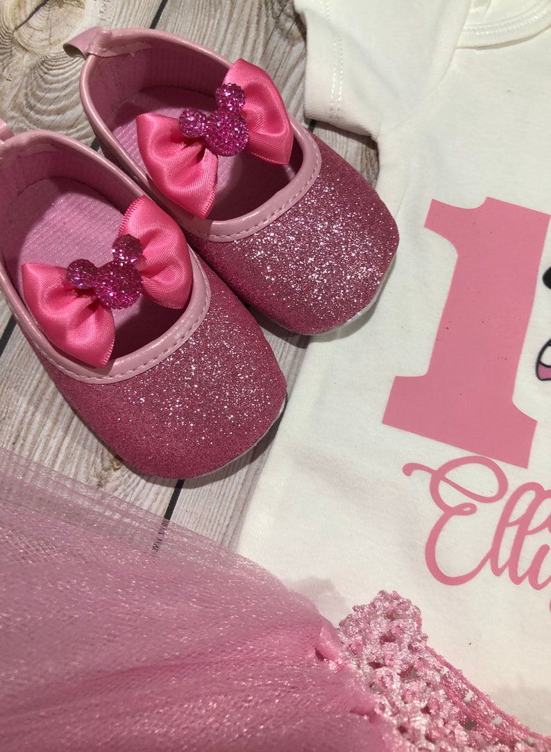 May include: Pink glitter baby shoes with a pink bow and a Minnie Mouse head design. The shoes are on a wooden surface.