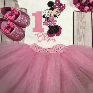 May include: Pink tutu with a white onesie featuring a pink number one and a Minnie Mouse graphic. The onesie has the text "Carter's 12m" and "Elison" on it. The outfit is completed with pink glitter shoes and a pink crown.