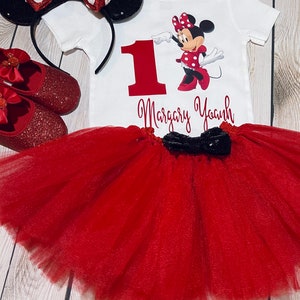 May include: A red and black Minnie Mouse themed first birthday outfit for a baby girl. The outfit includes a white bodysuit with a red number one, a red tulle tutu, a red headband with black ears and a red bow, and red glitter shoes with a red bow.