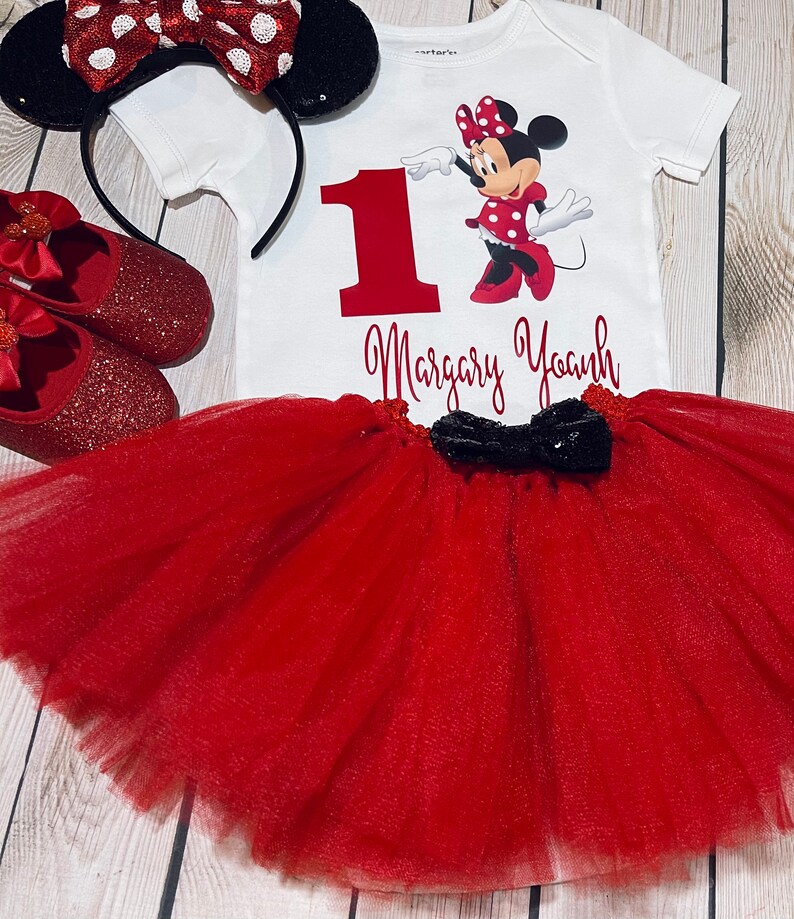 May include: A red and black Minnie Mouse themed first birthday outfit for a baby girl. The outfit includes a white bodysuit with a red number one, a red tulle tutu, a red headband with black ears and a red bow, and red glitter shoes with a red bow.