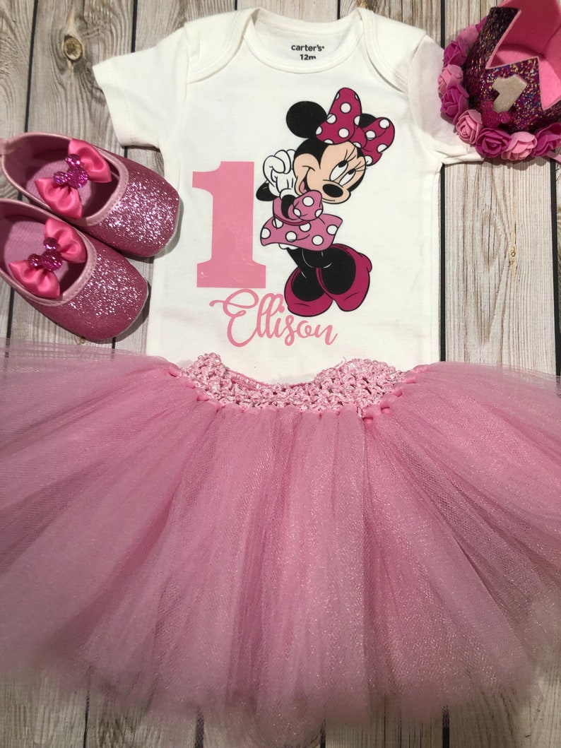 May include: Pink tulle skirt with a white bodysuit featuring a pink number one and a Minnie Mouse graphic. The bodysuit has the text "Carter's 12m" and "Ellison" printed on it. The outfit is completed with pink glitter shoes and a pink crown.