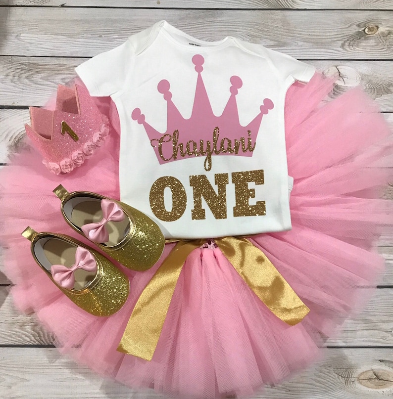 May include: Pink and gold princess-themed baby girl outfit with a crown, tutu, and shoes. The shirt says "Chaylani ONE" in gold glitter.