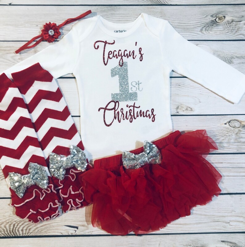 May include: A festive baby outfit set featuring a white long-sleeve bodysuit with the text "Teagan's 1st Christmas" in glittery red and silver. The set includes a red tutu skirt, red and white chevron leg warmers, and a red headband.