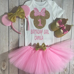 May include: A pink and gold Minnie Mouse themed first birthday outfit for a baby girl. The outfit includes a white bodysuit with a gold glitter Minnie Mouse head and the number 1, a pink tulle tutu with a gold bow, a pink and gold headband with ears, and gold glitter shoes with pink bows.