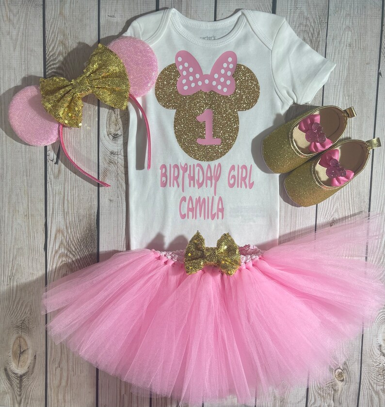 May include: A pink and gold Minnie Mouse themed first birthday outfit for a baby girl. The outfit includes a white bodysuit with a gold glitter Minnie Mouse head and the number 1, a pink tulle tutu with a gold bow, a pink and gold headband with ears, and gold glitter shoes with pink bows.