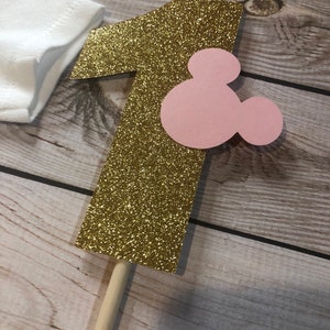 Puede incluir: N&uacute;mero uno de purpurina dorada con una cabeza de Mickey Mouse rosa unida. El n&uacute;mero uno est&aacute; en un palo de madera.