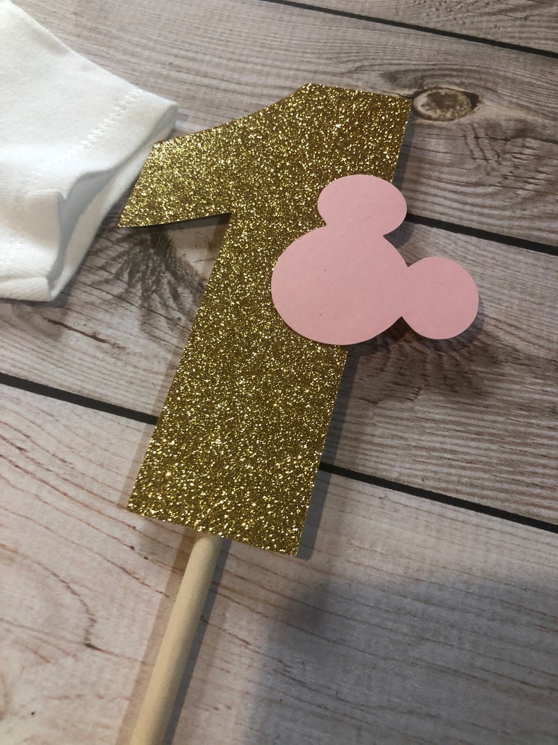 Puede incluir: N&uacute;mero uno de purpurina dorada con una cabeza de Mickey Mouse rosa unida. El n&uacute;mero uno est&aacute; en un palo de madera.