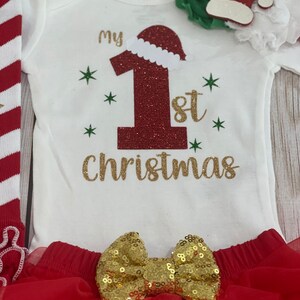 Pu&ograve; includere: Completo natalizio per neonati, composto da un body bianco con la scritta "my 1st Christmas" in glitter dorati, un grande numero 1 rosso glitterato con cappello da Babbo Natale, una gonna tutu rossa e un fiocco dorato.