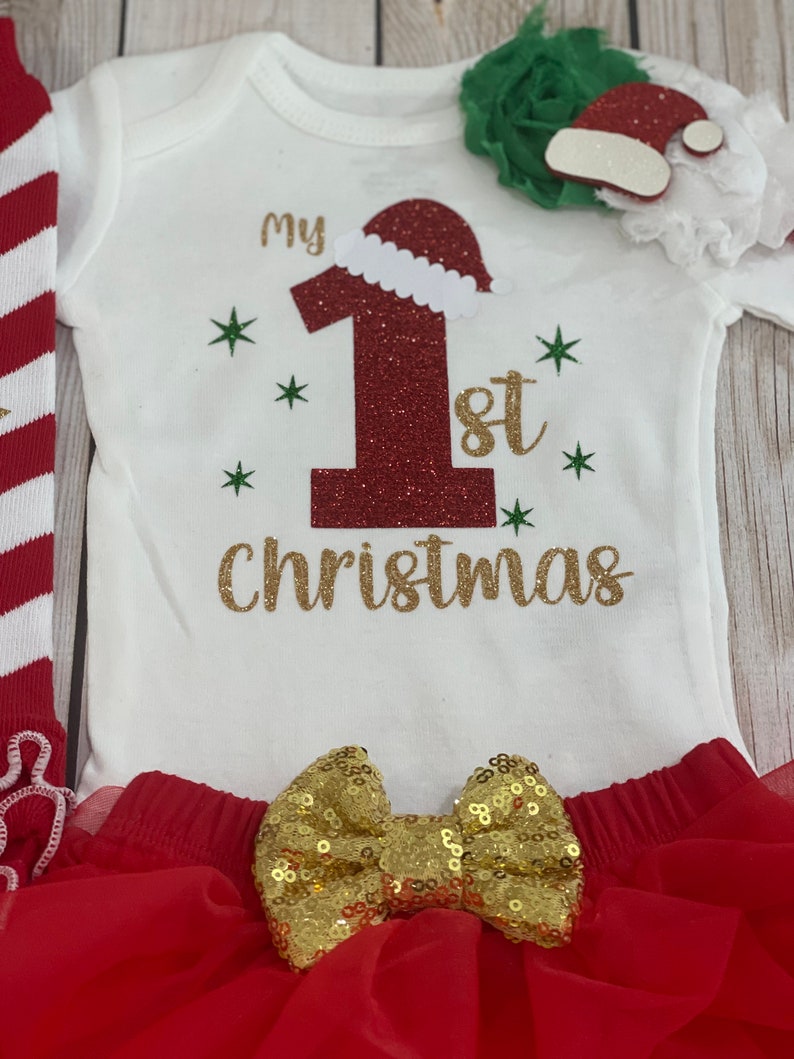 Pu&ograve; includere: Completo natalizio per neonati, composto da un body bianco con la scritta "my 1st Christmas" in glitter dorati, un grande numero 1 rosso glitterato con cappello da Babbo Natale, una gonna tutu rossa e un fiocco dorato.