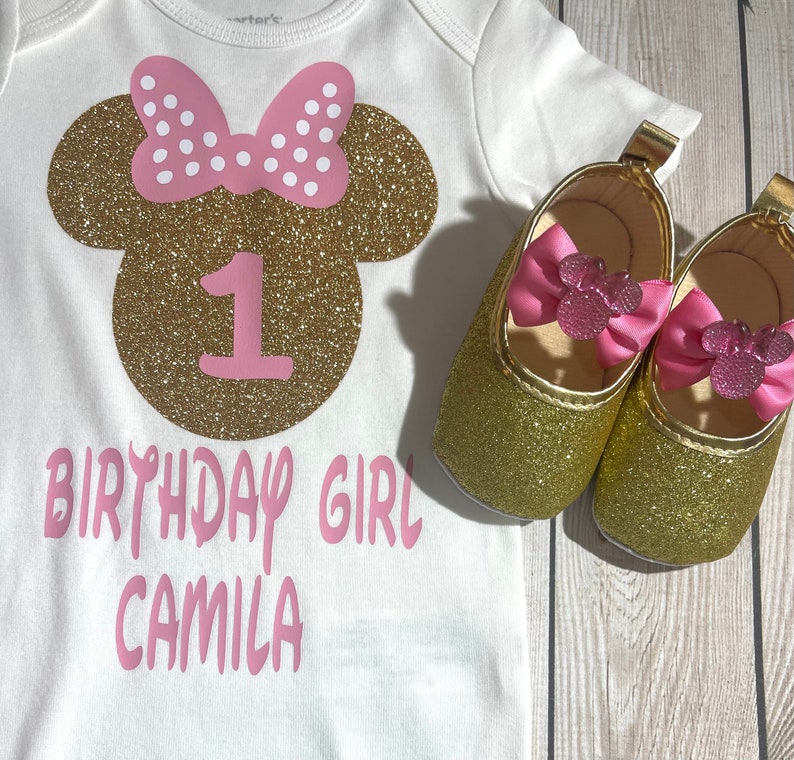 May include: A white onesie with a gold glitter Minnie Mouse head with pink polka dot bow and the number 1 in pink. The text "Birthday Girl Camila" is written in pink below the Minnie Mouse head. Two gold glitter shoes with pink bows are on a wooden surface.
