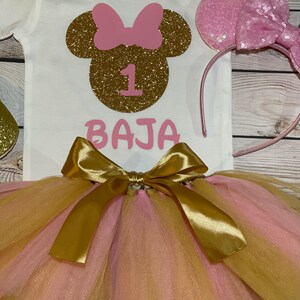 May include: A white onesie with a gold glitter Minnie Mouse head, a pink bow, and the number 1. The onesie also has the name "BAJA" written in pink letters. A pink tulle tutu with a gold satin bow is included. A pink headband with a pink bow and sequins is also included.