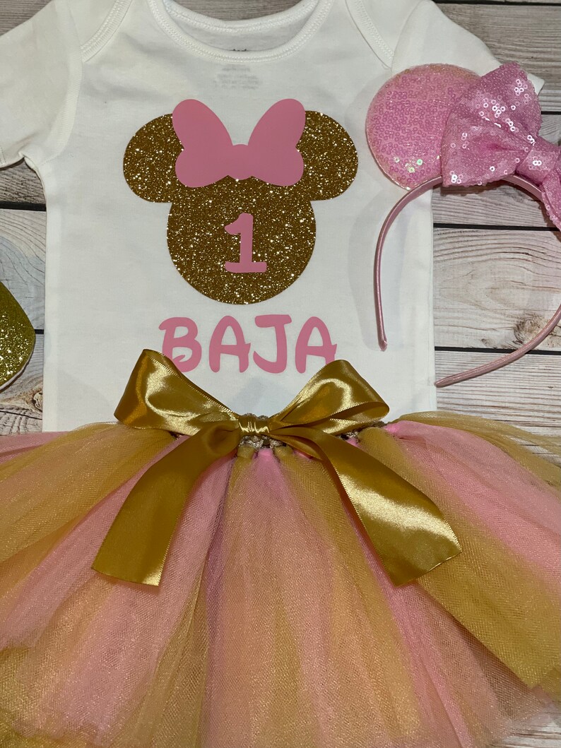 May include: A white onesie with a gold glitter Minnie Mouse head, a pink bow, and the number 1. The onesie also has the name "BAJA" written in pink letters. A pink tulle tutu with a gold satin bow is included. A pink headband with a pink bow and sequins is also included.