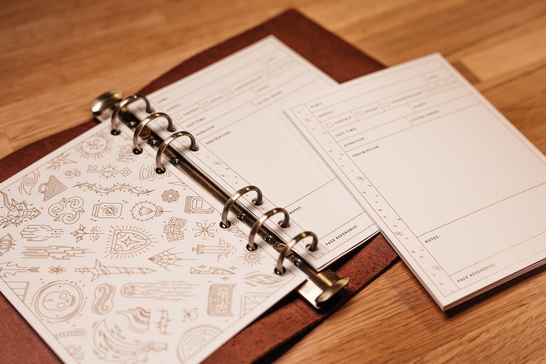 K&ouml;nnte beinhalten: Ein braunes, ledergebundenes Journal mit goldenen Ringen und einer wei&szlig;en Seite mit goldenen Tintenzeichnungen verschiedener Symbole. Die Seite ist auf eine leere wei&szlig;e Seite mit einem Abschnitt f&uuml;r Notizen und einer Seitenreferenz ge&ouml;ffnet.