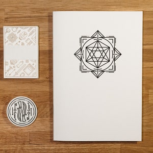 May include: A white notebook with a geometric design printed on the front cover. The design is black and features a square with a star inside. The notebook is on a wooden surface with two smaller white cards with geometric designs.