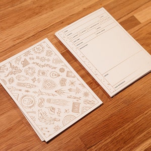 K&ouml;nnte beinhalten: Zwei wei&szlig;e Papiernotizb&uuml;cher, eines mit einer leeren Seite und das andere mit einem Muster aus verschiedenen Symbolen und Designs in goldener Tinte.