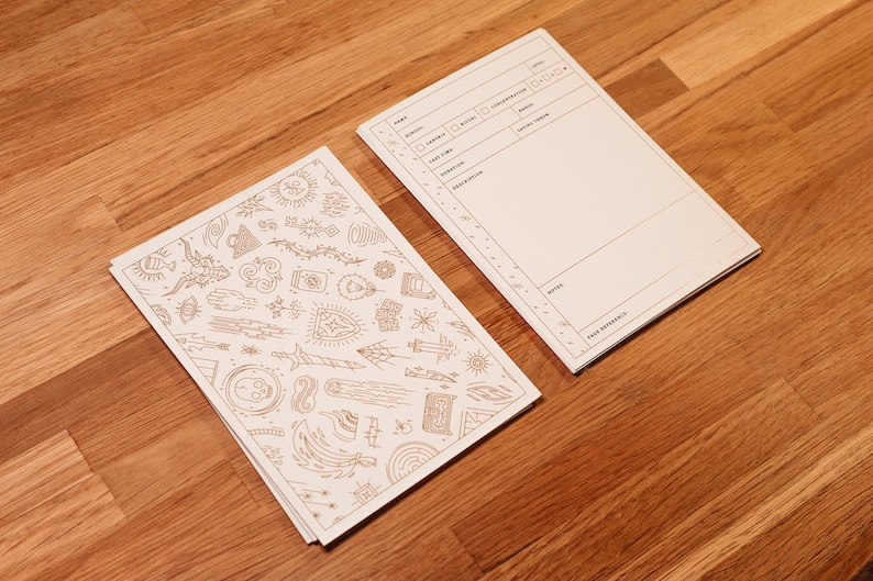 K&ouml;nnte beinhalten: Zwei wei&szlig;e Papiernotizb&uuml;cher, eines mit einer leeren Seite und das andere mit einem Muster aus verschiedenen Symbolen und Designs in goldener Tinte.