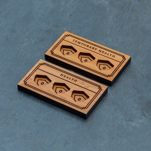 May include: Two wooden health trackers with engraved slots for tokens. The top tracker says "Temporary Health" and the bottom tracker says "Health".