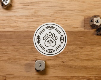 May include: A white round sticker with a black paw print design. The paw print has a sun rising above it. The sticker is surrounded by a black circle with leaves and wavy lines.