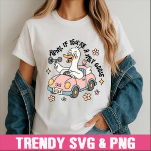 Peut inclure: T-shirt blanc avec un dessin d'oie de dessin anim&eacute; conduisant une voiture rose et klaxonnant. Le texte "Honk if you're a silly goose" est au-dessus de l'oie, avec le mot "Silly" sur la plaque d'immatriculation de la voiture. Des graphiques floraux et en forme d'&eacute;toiles entourent le dessin.