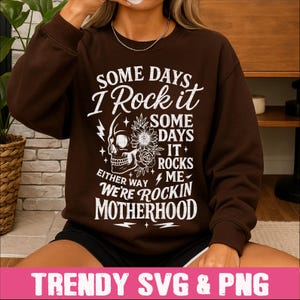 May include: A brown sweatshirt with a white graphic design that says "Some days I rock it, some days it rocks me, either way we're rockin motherhood". The graphic also includes a skull with flowers.