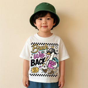 May include: A white t-shirt with a cartoon goose wearing sunglasses, a hat, and sneakers. The shirt has the words "Bruh We Back" in a colorful design. The goose is carrying a book that says "Day One Drama". The child is wearing a green bucket hat.