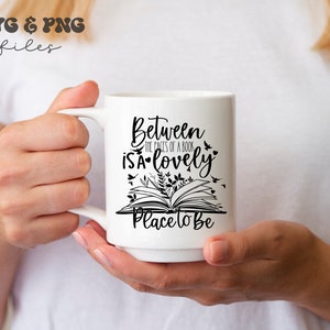 May include: A white ceramic mug featuring black text that reads Between the pages of a book is a lovely place to be, accompanied by a graphic illustration of an open book with small leaves, birds, and hearts surrounding the printed lettering.