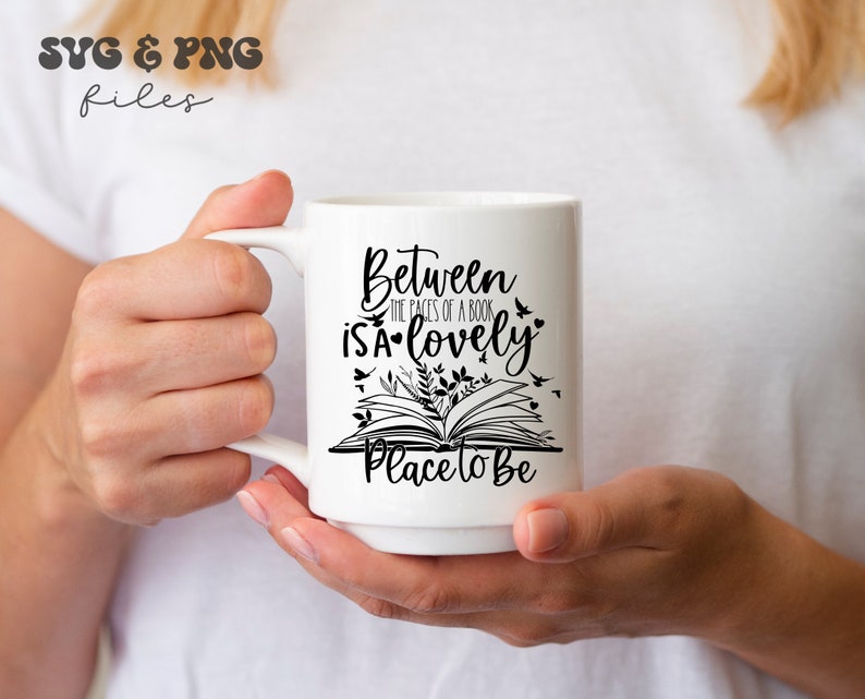 May include: A white ceramic mug featuring black text that reads Between the pages of a book is a lovely place to be, accompanied by a graphic illustration of an open book with small leaves, birds, and hearts surrounding the printed lettering.