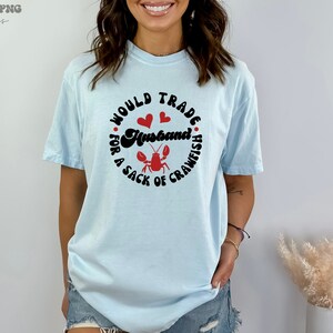 May include: A light blue t-shirt with the text "Would trade a husband for a sack of crawfish" in black with red hearts and a red crawfish illustration.