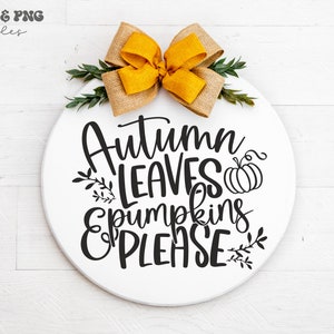 May include: A circular white wooden sign featuring the black text Autumn LEAVES & PUMPKINS PLEASE with a small pumpkin and leaf illustrations. A large mustard yellow burlap bow and green foliage are attached to the top of the round sign.