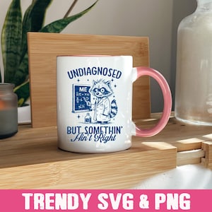 May include: White ceramic mug with a pink handle. The mug has a blue graphic of a raccoon wearing a lab coat and holding a test tube. The text on the mug reads "Undiagnosed But Somethin' Ain't Right".