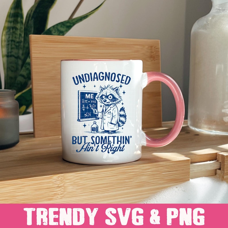 May include: White ceramic mug with a pink handle. The mug has a blue graphic of a raccoon wearing a lab coat and holding a test tube. The text on the mug reads "Undiagnosed But Somethin' Ain't Right".