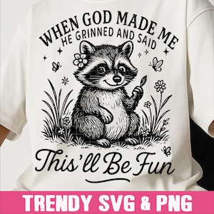 Op de afbeelding: Wit T-shirt met een zwart-witte illustratie van een lachende wasbeer die een blad vasthoudt, omringd door bloemen en gras. De tekst luidt "WHEN GOD MADE ME HE GRINNED AND SAID This'll Be Fun".